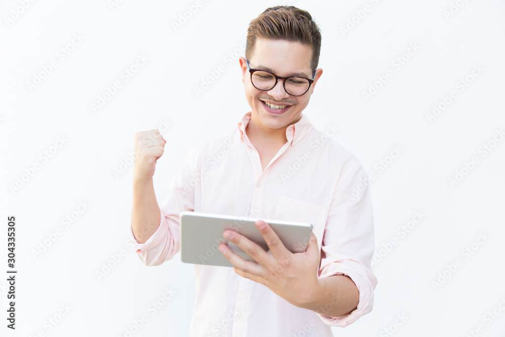 Happy excited tablet user celebrating success and making winner gesture. Young man in glasses standing isolated over white background. Communication or good news concept Happy excited tablet user celebrating success and making winner gesture. Young man in glasses standing isolated over white background. Communication or good news concept