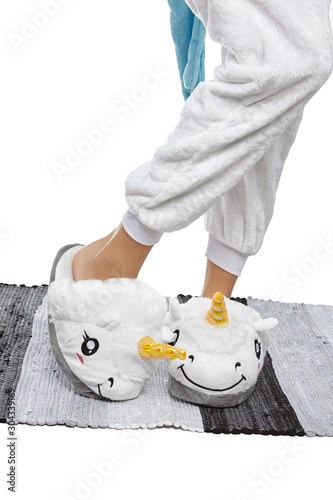 Close-up shot of female legs in white velour trousers and plush house slippers made in the form of white smiling unicorn. The girl is standing on the striped gray and white carpet.
