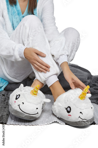 Cropped medium shot of a lady in white velour pyjamas and plush house slippers made in the form of white smiling unicorn. The girl is sitting on the striped carpet and a gray plaid.