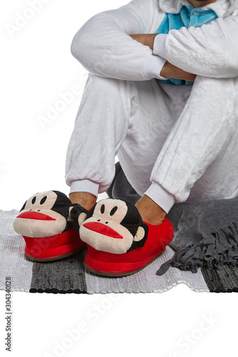 Cropped medium shot of a man in white velour pyjamas and red plush house slippers made in the form of monkey head. The man is sitting on the striped carpet and a gray plaid.