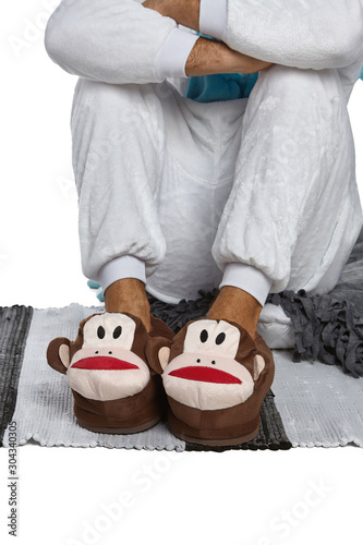 Close-up shot of male legs in white velour trousers and brown plush house slippers made in the form of monkey head. The man is standing on the striped gray and white carpet.