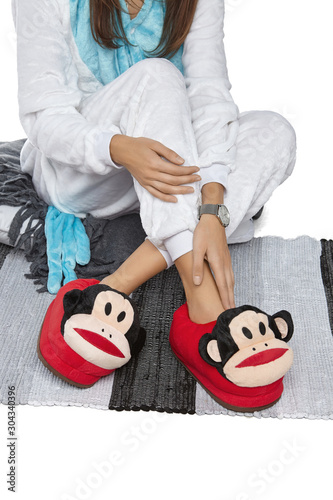 Cropped medium shot of a lady in white velour pyjamas and red plush house slippers made in the form of monkey head. The girl is sitting on the striped carpet and a gray plaid.