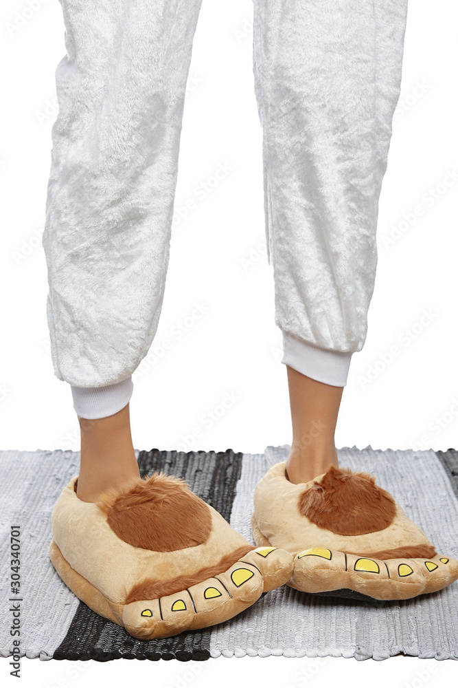 Close-up shot of female legs in white velour trousers and brown plush house  slippers made as shaggy hobbit feet. The girl is standing on the striped  gray and white carpet. Photos