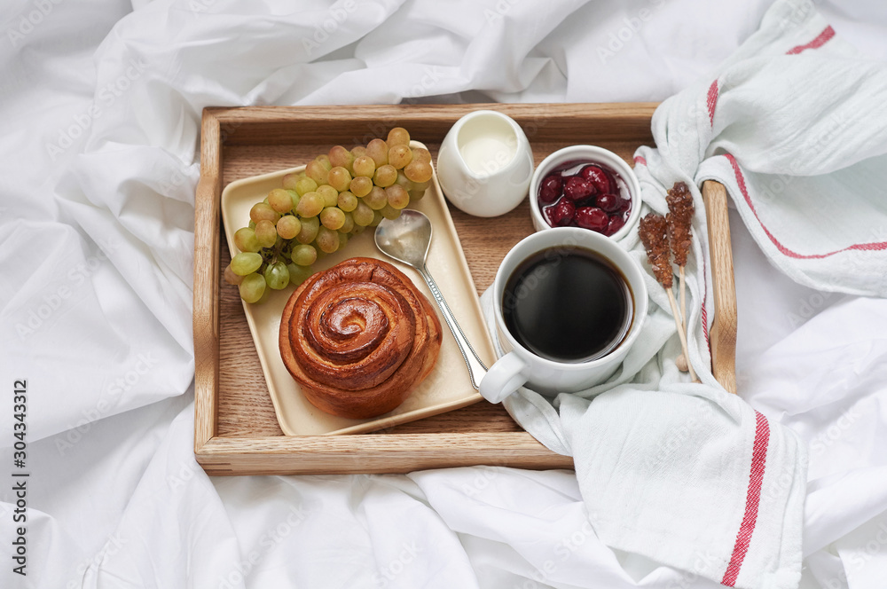 Breakfast on white bed sheets in hotel room. Close up of wooden tray with coffee, bun, jam and grape