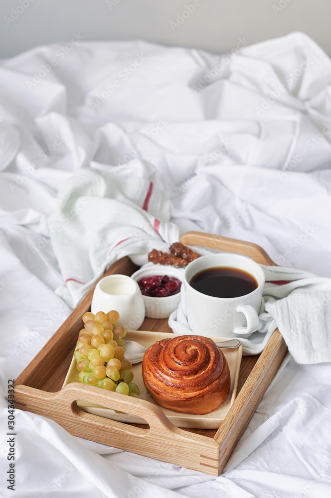 Breakfast on white bed sheets in hotel room. Close up of wooden tray with coffee, bun, jam and grape