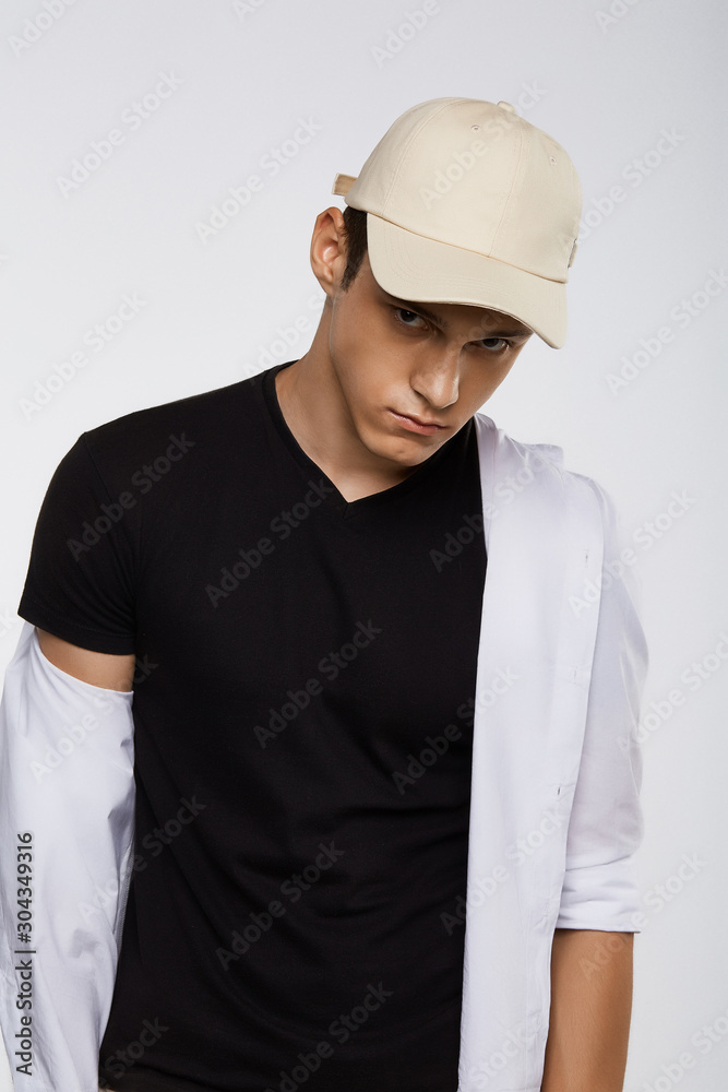 Cropped front view shot of a dark-haired man, wearing beige baseball cap, black t-shirt and white shirt. He is posing on grey background and looking straight. 