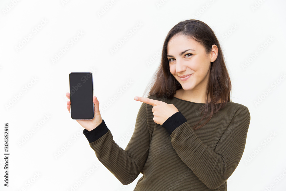 Happy joyful cellphone user pointing at blank screen. Young woman in casual with mobile phone standing isolated over white background. Application or software advertising concept Happy joyful cellphone user pointing at blank screen. Young woman in casual with mobile phone standing isolated over white background. Application or software advertising concept