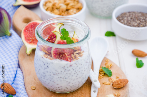 Chia pudding with yogurt, homemade granola, fresh figs and honey in a glass jar on a board on a white wooden background.