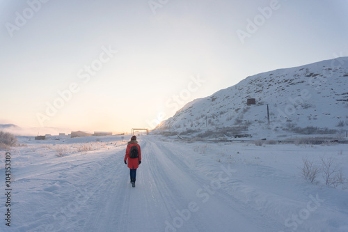 a lonely girl in a bright jacket walks along a white snowy road through the tundra beyond the Arctic Circle on a frosty clear day