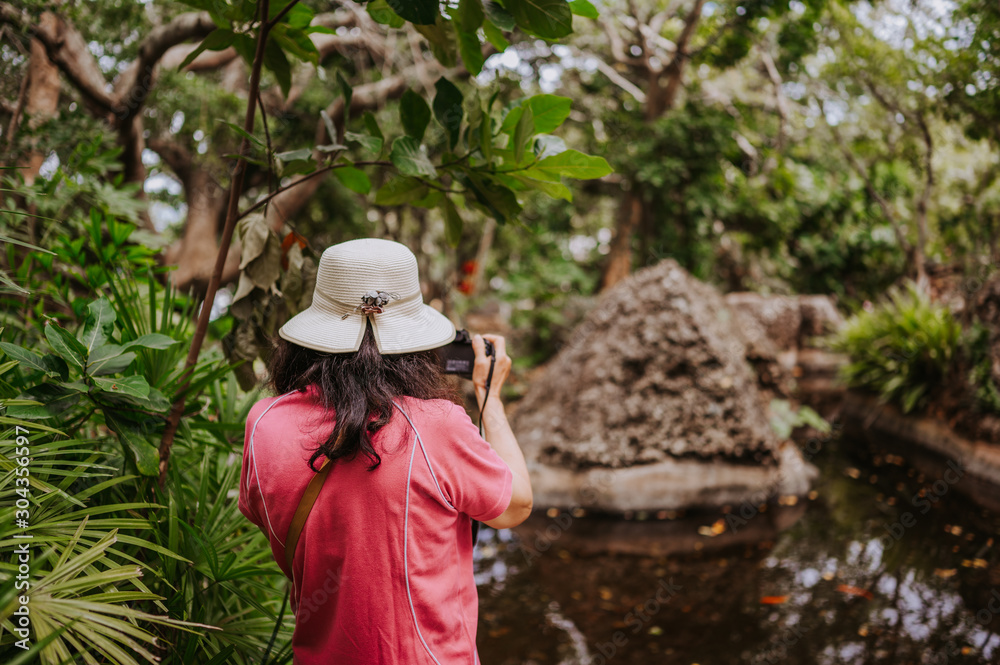 Asian tourist woman in national hat taking photos at natural green park with tropical plants and palm trees. Travel Asia tourism
