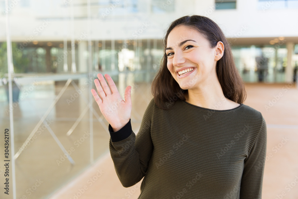 Friendly happy Latin woman waving hello. Young woman in casual posing indoors with glass wall interior in background. Greeting or communication concept