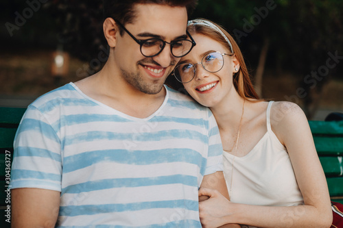 Portrait of a beautiful couple embracing while sitting on a bench outdoor while woman with red hair and freckles is looking at hims smiling leaning her head. photo