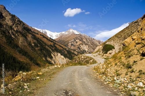 A dirt road running along the canyon, a picturesque mountain autumn landscape on a bright sunny day, in the center of the photo is a mountain peak with a small snow cap, Kyrgyzstan
