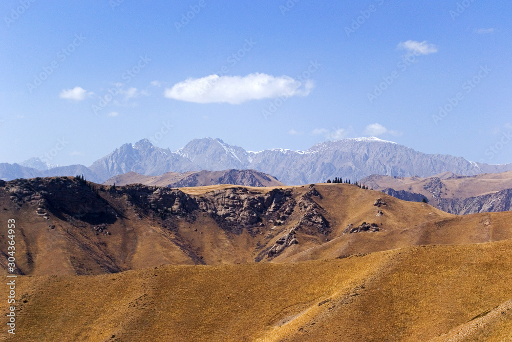 Mountains of Kyrgyzstan on a bright sunny day. The mountains like a layered cake are located one after another. Near without stones, like a hill. Far high mountain with snow peaks