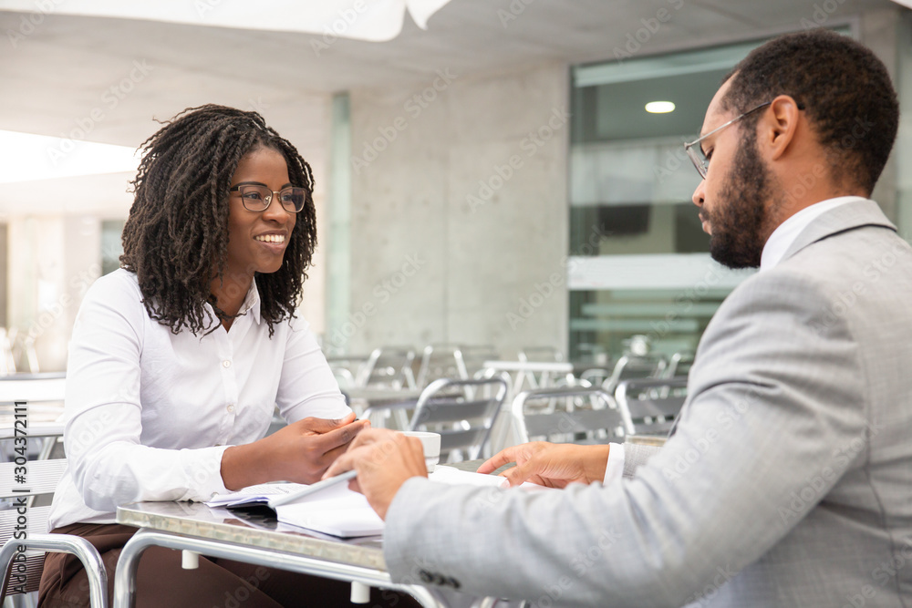 Smiling African American woman in eyeglasses looking at colleague. Two businesspeople working at outdoor cafe. Business concept Smiling African American woman in eyeglasses looking at colleague. Two businesspeople working at outdoor cafe. Business concept