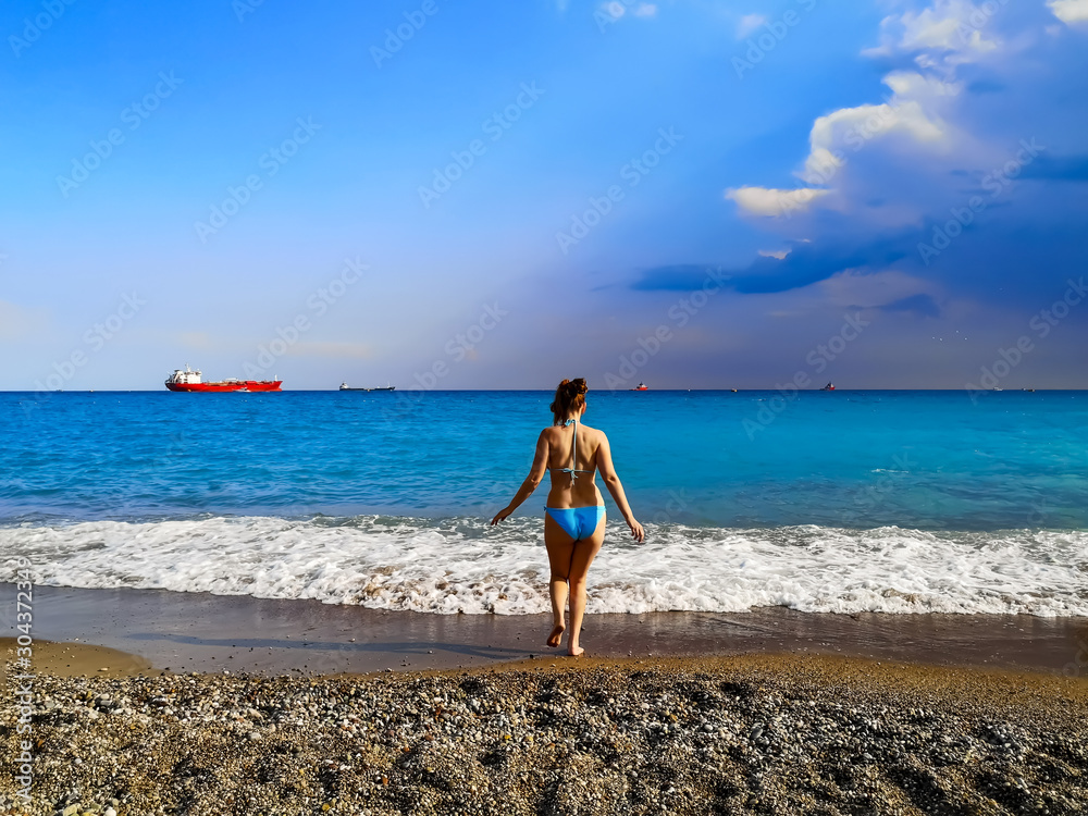 Obraz premium Seascape with many ships on the horizon and one woman on the beach. A young adult woman in a swimsuit with a slim figure enters the sea water with white foam
