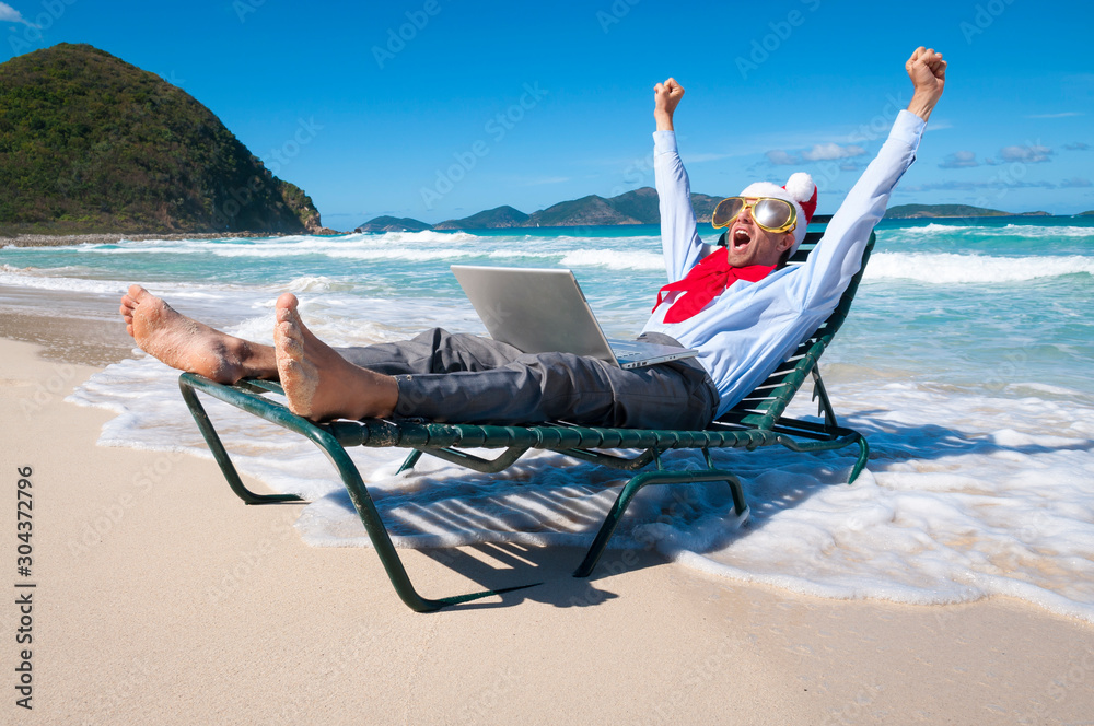 Excited Santa businessman celebrating Christmas shopping success with his laptop on a tropical beach