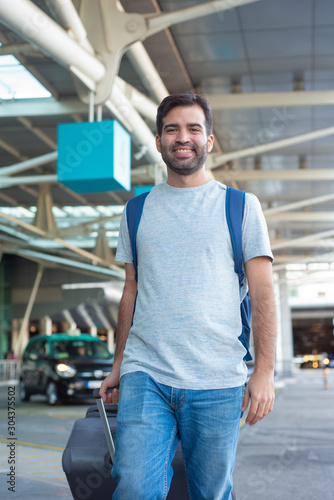 Cheerful young man walking near airport. Smiling young traveler looking at camera. Travel concept