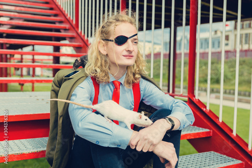 A theatrically dressed pseudo soldier or pirate with a blindfold in one eye sits on the stairs. Young man with a viking haircut. A white rat runs along his arm.	 photo