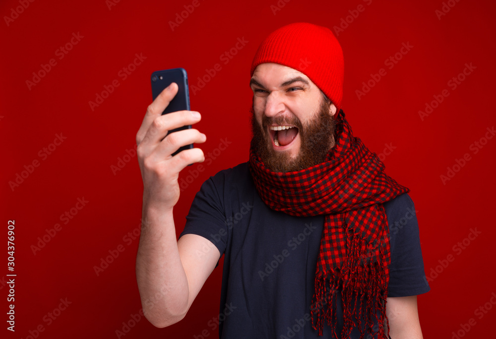 Photo of screaming bearded guy, wearing red hat and scarf, using mobile phone Photo of screaming bearded guy, wearing red hat and scarf, using mobile phone