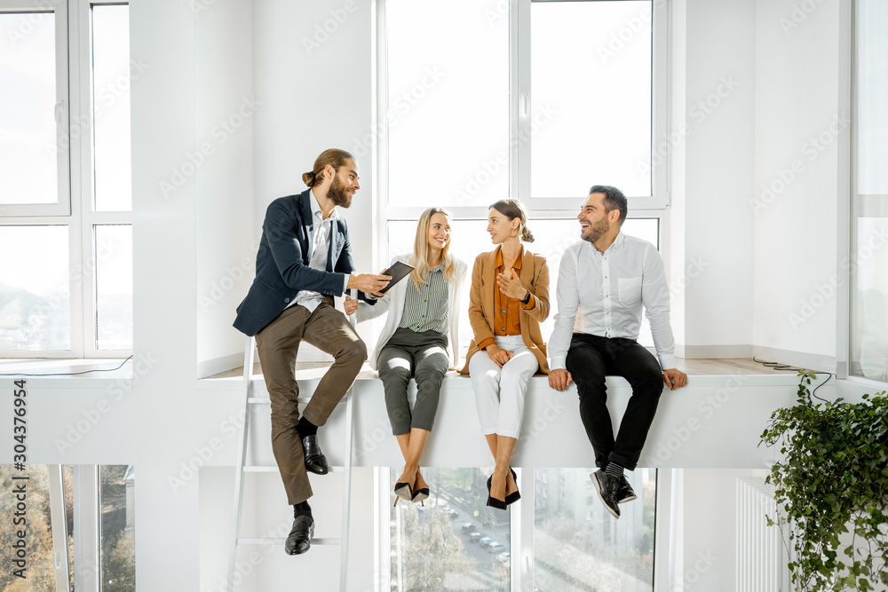Fototapeta premium Group of a young creative office employees having some informal discussion, while standing in the two-level bright meeting room with large windows