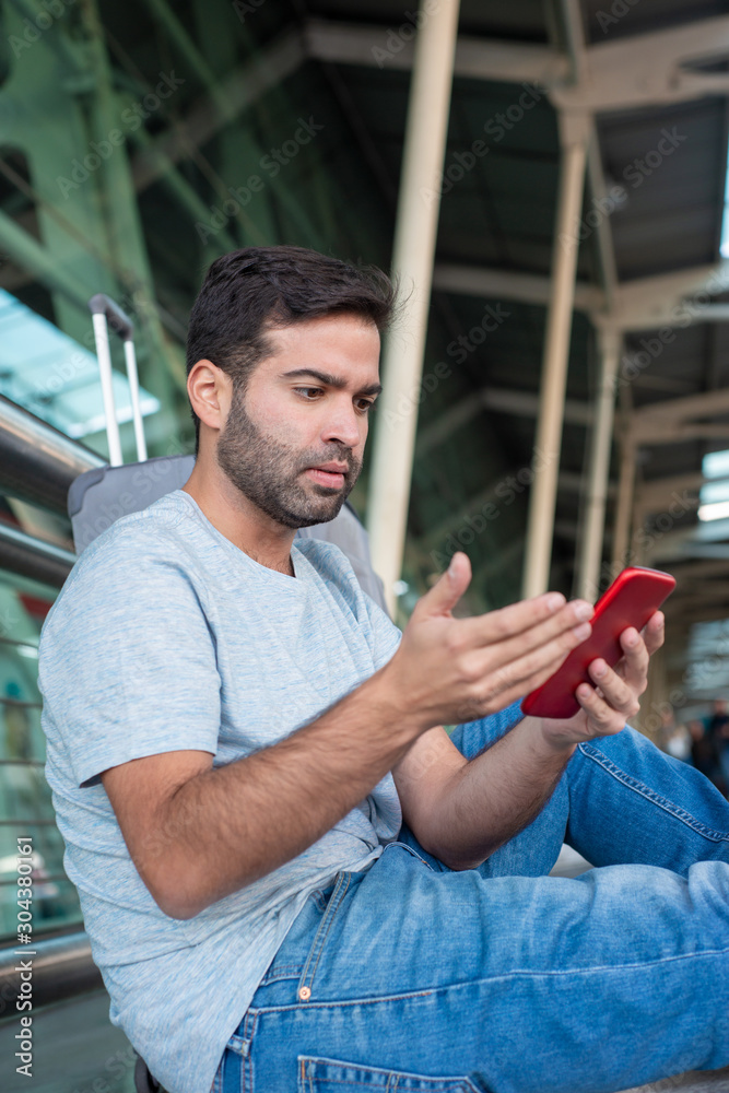 Disgruntled young traveler looking at smartphone. Dissatisfied Hispanic man sitting on floor and using phone. Travel concept - obrazy, fototapety, plakaty 