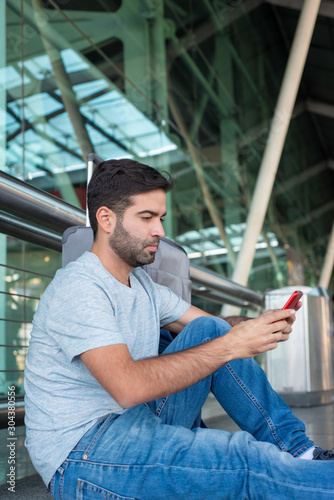 Thoughtful young man sitting on floor and looking at phone. Focused handsome bearded Hispanic guy using smartphone at airport. Travel concept