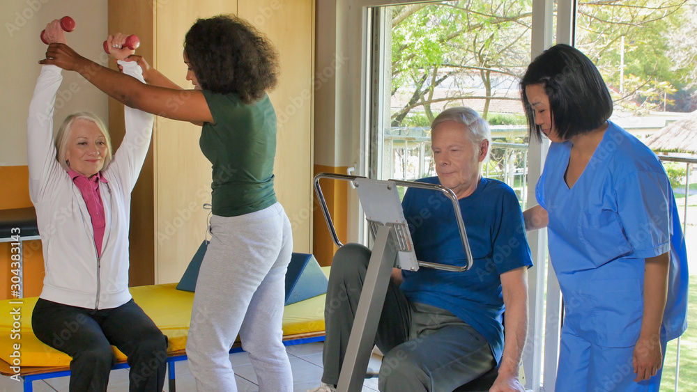 Rehabilitation concept. Elderly retired couple training in a gym supervised by expert african and asian female trainers