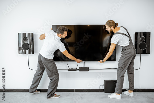 Photos Two professional workmen in workwear installing a large TV monitor and audio system in the white living room
