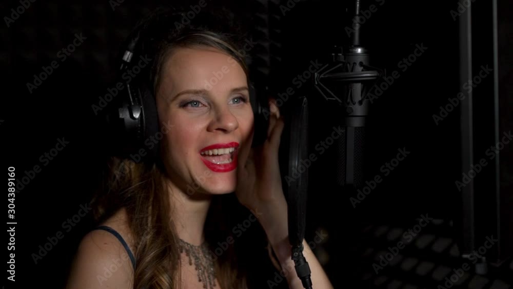Close-up portrait of a female singer singing in headphones into a microphone in the dark, slow motion Close-up portrait of a female singer singing in headphones into a microphone in the dark, slow motion