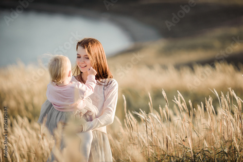 mother with red hair in pastel clothes looks on a blonde daughter in her arms in a gray tulle skirt in a wheat field, in a sunny warm sunset light