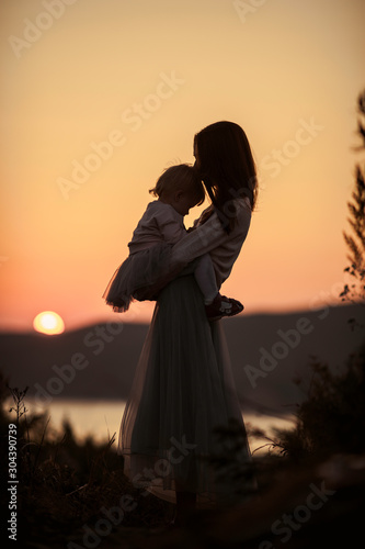 Silhouettes of mother with a baby in her arms on nature with a view of the sun setting in the water, evening at golden hour