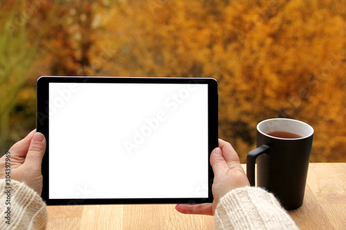 girl holds horizontally tablet PC with blank white screen, black mug with tea on the terrace over autumn yellow and orange trees, the concept of a cozy autumn in nature