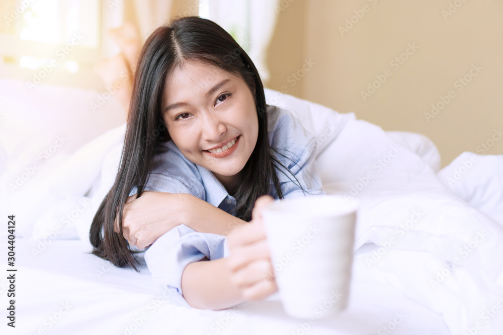 Gorgeous smile young woman with blanket holding white cup of hot coffee or tea while lying on the bed in the morning after wake up in the bedroom. Gorgeous smile young woman with blanket holding white cup of hot coffee or tea while lying on the bed in the morning after wake up in the bedroom.