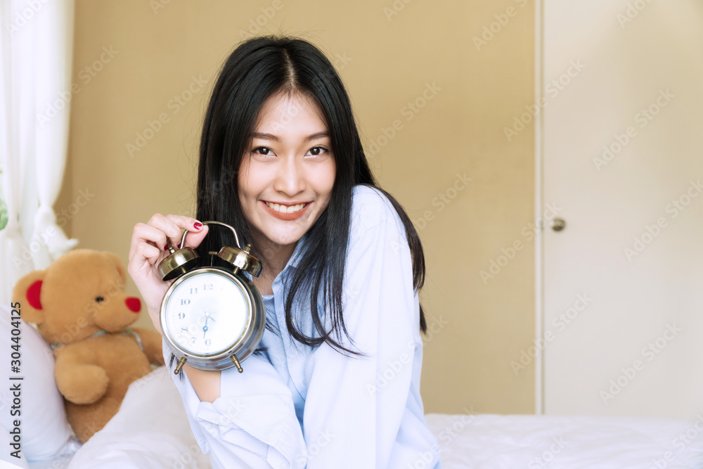 Smiling young woman with teddy bear holding alarm clock sitting on the white bed while looking at camera. Cheerful girl waking up in the morning showing alarm clock in her hand. Lifestyle concept. Smiling young woman with teddy bear holding alarm clock sitting on the white bed while looking at camera. Cheerful girl waking up in the morning showing alarm clock in her hand. Lifestyle concept.