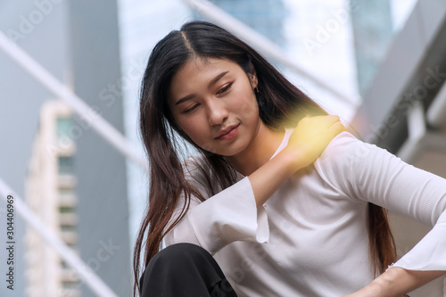 Upset young woman with shoulder pain or stiffness while sitting on city background. Businesswoman overwork tired while her hand touch neck problem stress after work office. Health care concept.