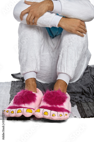 Cropped medium shot of a man in white velour pyjamas and pink plush house slippers made as shaggy hobbit feet. The man is sitting on the striped carpet and a gray plaid.