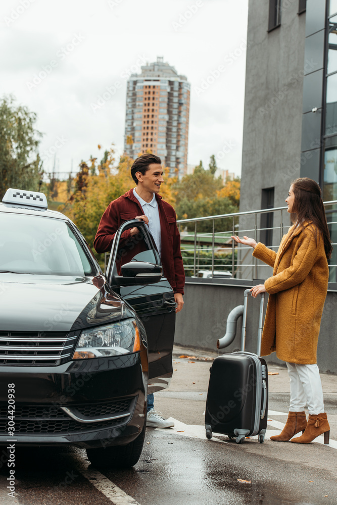 Fototapeta premium Woman with suitcase talking with taxi driver beside car