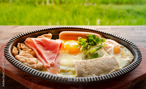 Breakfast Asian style on hot plate. KAI KATA  that made from fried egg with pepper and some meat such as chinease sausage , pork on a wooden table Amid the view of the rice fields backgroud. photo