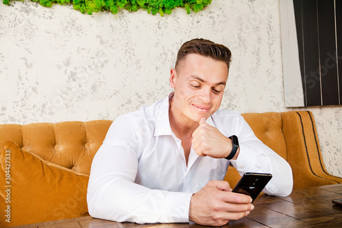 Inflated guy in shirt sits in cafe at a table with phone in his hand.