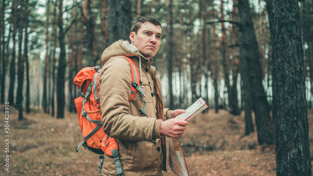 A traveling man with map in woods. A portrait of the handsome man with a backpack, standing near a tree in a cold weather. A traveling man with map in woods. A portrait of the handsome man with a backpack, standing near a tree in a cold weather.
