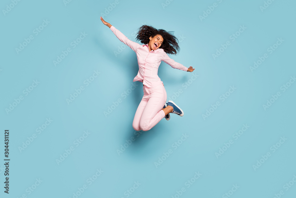 Full length photo of amazing dark skin lady jumping high spread hands sides spending best free time wear pink shirt trousers sneakers isolated blue color background