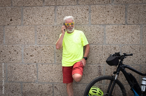 an old man dressed in red and yellow colors leaning against the concrete wall talking on the phone. Electric bicycle near him