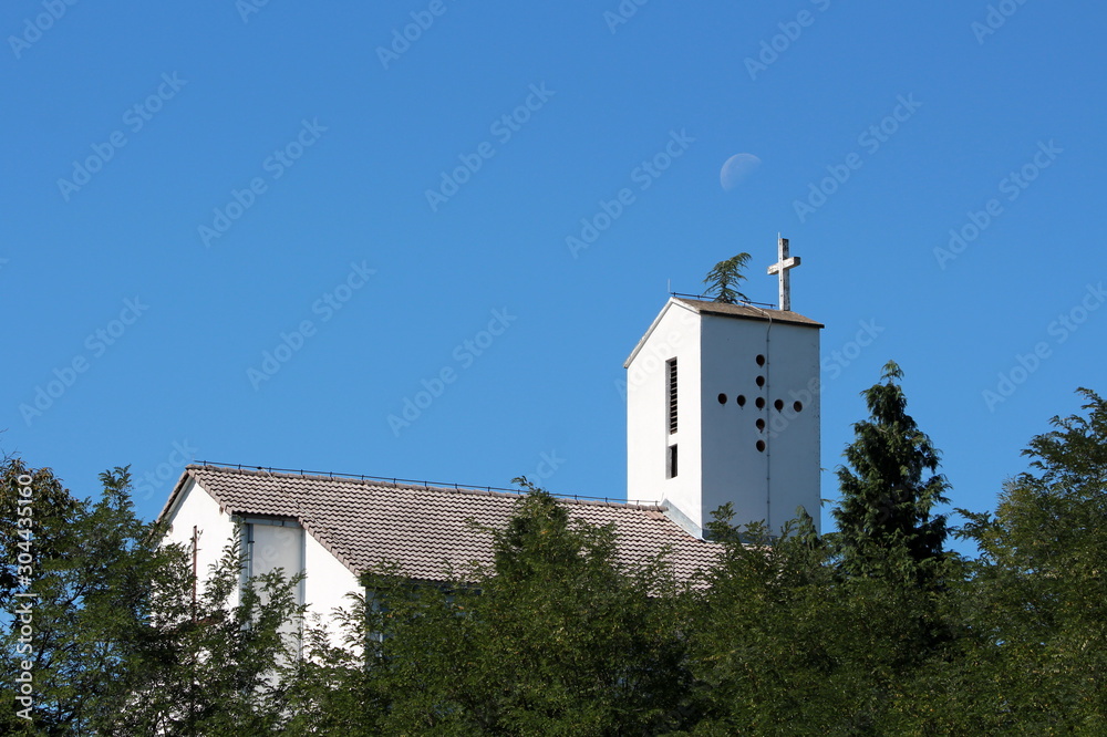 Narrow white catholic church with concrete cross on top completely surrounded with dense trees and forest vegetation on clear blue sky with barely visible moon in background on warm summer day
