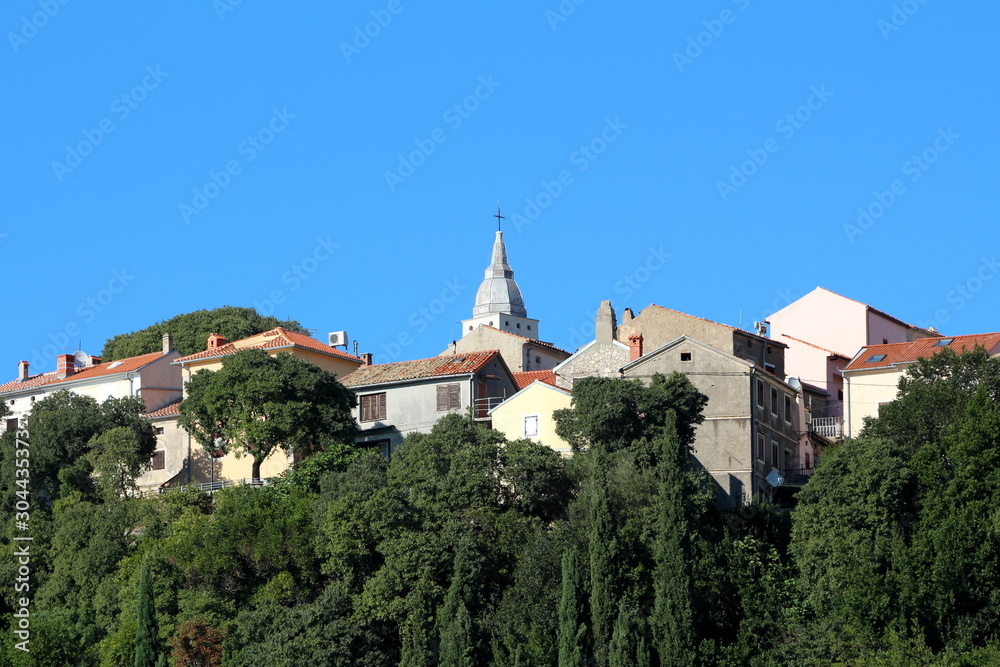 Old Mediterranean town filled with densely built stone family houses around local catholic church rising high above small hill surrounded with dense forest and clear blue sky in background