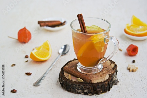 Apple cider with spices. Alcoholic or non-alcoholic hot drink made from fresh unfiltered apple juice with spices, apple slices and orange in a glass mug on a light concrete background.