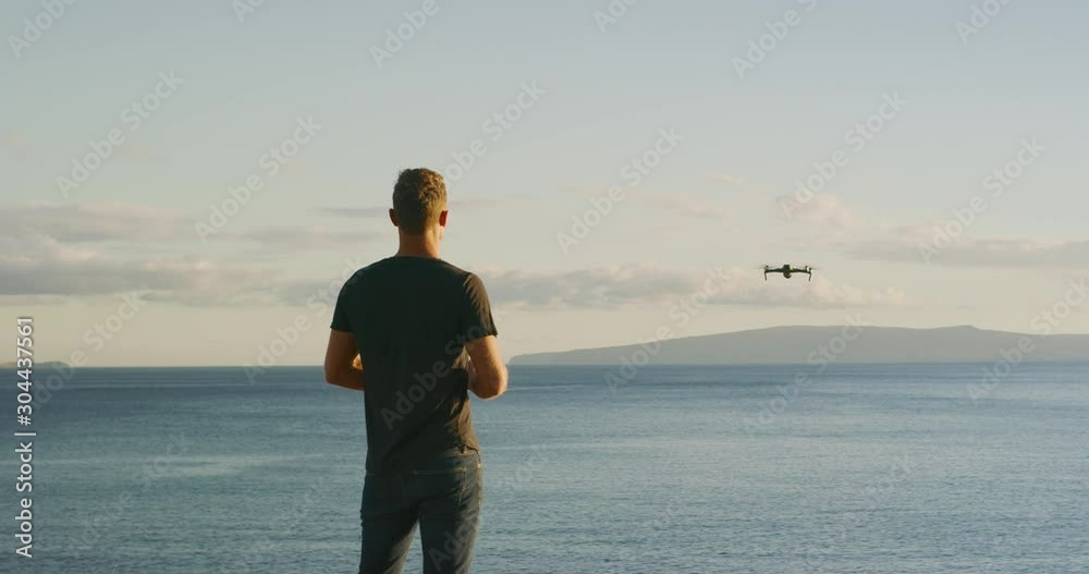 A young man flying his new quadcopter drone out over the ocean at sunset, drone pilot flying at sunset