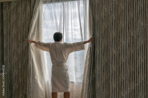 Behide asian man in bathrobe suit open curtain to see view outside in morning, Travel and Holiday concept