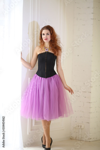 beautiful red-haired girl in a purple tulle skirt and black corset is standing near the floor window