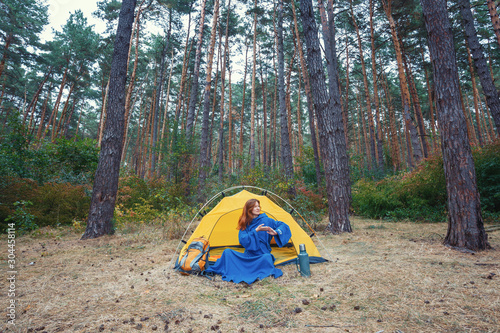 Young pretty redhead girl tourist in blue robe stretching hands after wake up, doing exercises, sitting in yellow tent, relaxing, enjoying nature in autumn forest. Travel weekend outdoors concept